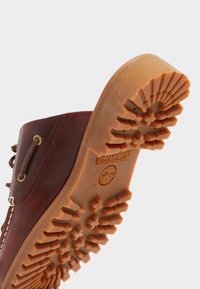 Bota de cuero marrón con una suela de goma texturizada. La suela presenta un patrón de huella distintivo y un logo circular. Se pueden ver las ojetes para los cordones.