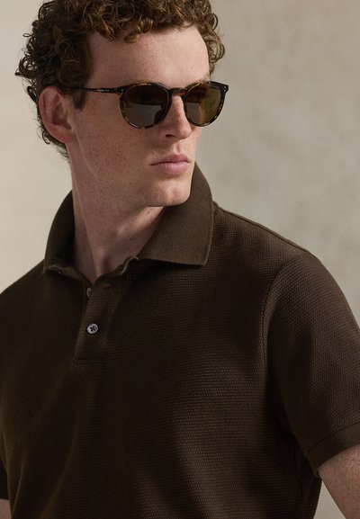 Brown textured polo shirt with a buttoned collar; wearing tortoiseshell sunglasses. Close-up view focusing on the shirt's weave and collar.