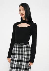 Μαύρη μπλούζα με μακριά μανίκια και κομμένη λαιμόκοψη, σε συνδυασμό με μια γκρι και μαύρη καρό φούστα που διαθέτει μια μαύρη ζώνη με κυκλικό κούμπωμα.