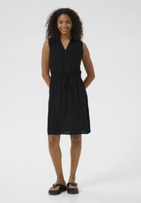 Schwarzes ärmelloses Kleid mit durchgehender Knopfleiste, verstellbarem Taillenbund und geradem Schnitt. Glatter Stoff, knielang, kombiniert mit schwarzen Sandalen.