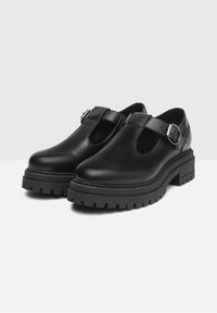 Scarpe Mary Jane in pelle nera con suole spesse e strap regolabili, esposte su una superficie bianca con sfondo grigio.