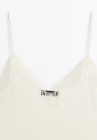 Débardeur en crème avec un décolleté en dentelle en V, des bretelles fines et une texture douce. Étiquette visible au centre, sous les détails en dentelle.