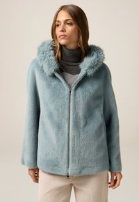 Giacca in pelliccia sintetica di colore azzurro chiaro con zip frontale e cappuccio soffice. Presenta maniche ampie ed è abbinata a un maglione a collo alto scuro.
