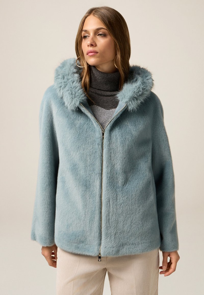 Giacca in pelliccia sintetica di colore azzurro chiaro con zip frontale e cappuccio soffice. Presenta maniche ampie ed è abbinata a un maglione a collo alto scuro.