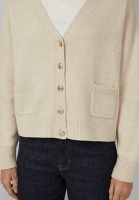 Beige gebreide cardigan met een diepe V-hals, vijf metalen knopen, geribde manchetten en twee voorzakken. Zachte textuur, casual pasvorm.