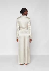 Silke pysjamassett i lys beige. Skjorten har krage og lange ermer, mens buksene har vide ben, noe som gir en flytende silhuett.