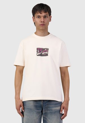 Jonge man die een wit T-shirt draagt met een kleine paarse en zwarte afbeelding in het midden en blauwe jeans, staand tegen een effen achtergrond.