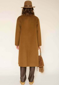 Manteau en laine marron à manches longues, coupe droite, avec un détail de couture au dos, accompagné d'un pantalon en cuir marron et d'un chapeau en feutre assorti.