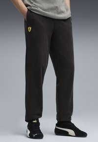 Person iført sorte Ferrari sweatpants og sorte Puma sneakers med hænderne i lommerne mod en grå baggrund.