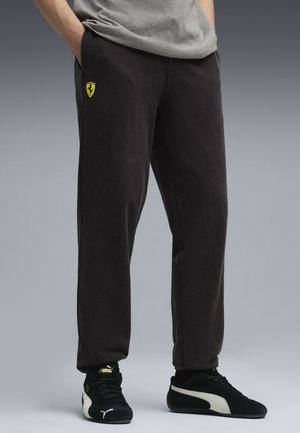 Person iført sorte Ferrari sweatpants og sorte Puma sneakers med hænderne i lommerne mod en grå baggrund.