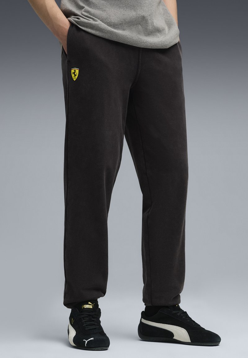 Person iført sorte Ferrari sweatpants og sorte Puma sneakers med hænderne i lommerne mod en grå baggrund.