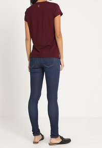 Burgundin lyhythihainen t-paita, jossa on logo takana, yhdistettynä tummansinisisiin slim fit -farkkuihin ja mustiin liukuhalkaisijakenkiin, esiteltynä takaa.