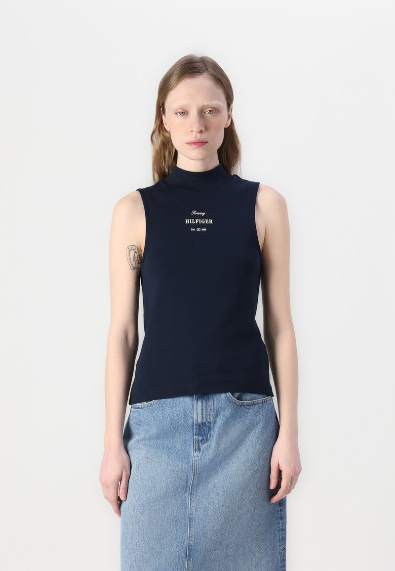 Jūras spēku bez piedurknēm augsts apkakles tops ar baltu "Tommy HILFIGER" logotipu. Gluda auduma, pieguļošs dizains, kombinēts ar gaiši zilu džinsu svārku.