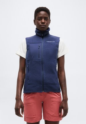 Άτομο που φοράει μπλε φλις γιλέκο πάνω από λευκό κοντομάνικο πουκάμισο και κοραλλί cargo σορτς, στέκεται μπροστά σε απλό ανοιχτό φόντο.