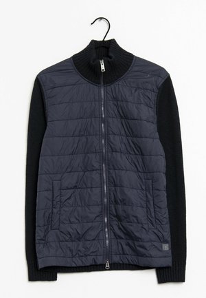 Czarna pikowana kamizelka na zamek z dzianinowymi, długimi rękawami i wysokim kołnierzem, wisząca na czarnym wieszaku na białym tle.