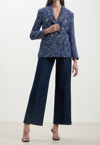 Femme portant un blazer croisé à carreaux bleus, un jean large foncé et des talons ouverts noirs, debout devant un fond uni.