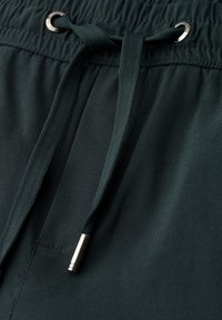 Donkergroene stoffen broek met een elastische tailleband en een zwart trekkoord gestrikt door metalen oogjes.