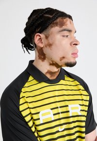 Sportisks polo krekls ar melniem un dzelteniem horizontāliem svītrainiem, ar melnu apkakli un baltu Nike logotipu priekšpusē.
