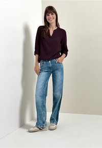 Bordeaux bluse med V-hals og opkrøllede ærmer, parret med lyseblå vide jeans og beige sneakers på en neutral baggrund.