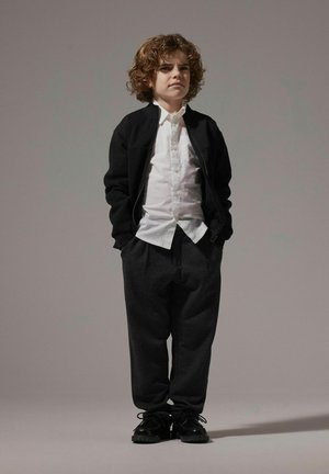 Veste zippée noire sur une chemise blanche à boutons, associée à un pantalon de survêtement gris foncé et des chaussures noires. Cheveux bouclés et arrière-plan neutre.