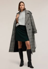 Langer karierter Mantel in Schwarz und Weiß über einem grauen Hoodie, kombiniert mit einem grünen Rock und knielangen schwarzen Stiefeln mit Absatz.