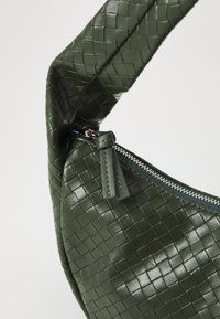 Green woven leather bag with a curved shape, featuring a zip closure and a textured surface. The strap is thick and softly structured.