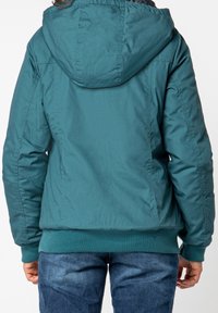 Veste isolée teal avec capuche, ourlet côtelé et texture lisse. Comprend des poches zippées et une coupe décontractée. Portée par-dessus un jean bleu.