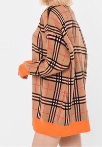 Femme portant un pull à carreaux beige avec des lignes noires, rouges, et des poignets et ourlets orange vif, vue de profil sur un fond blanc.