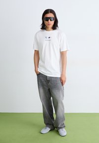 Hvid bomulds t-shirt med BMW Motorsport logo, parret med falmede grå brede jeans og hvide sneakers. Modellen bærer store sorte solbriller.