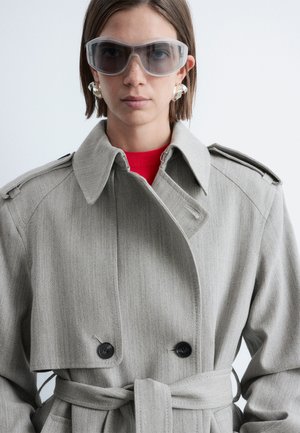 Personne portant un trench-coat gris surdimensionné, de larges lunettes de soleil teintées, des boucles d'oreilles argentées en forme d'anneau et un haut rouge, faisant face à l'avant sur un fond uni.