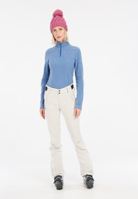 Pullover in pile blu con collo a zip, pantaloni da sci crema con tasche e dettagli neri a contrasto, abbinati a un berretto di lana rosa.