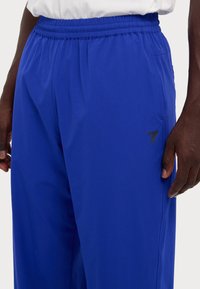 Pantalons de sport bleus en tissu léger, avec une taille élastique, des poches latérales et un petit logo noir sur la cuisse.
