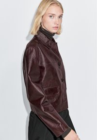 Vinrød læderjakke med krave, knaplukning foran og sidelomme. Glat tekstur, kort design. Modellen bærer en mørk rullekrave.