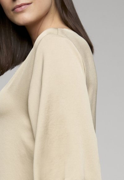 Femme aux cheveux bruns raides portant un haut beige à manches longues et amples, sur un fond gris uni.