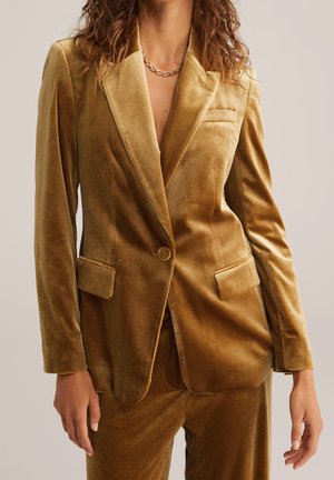 Kvinde iført en sennepsgul fløjlsblazer med en enkelt knap og matchende bukser, accessoriseret med en guldkæde halskæde.