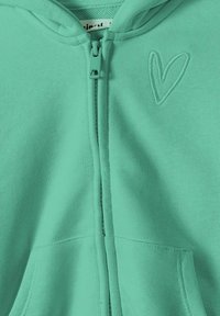 Sudadera con cremallera de color verde menta, hecha de un tejido suave, con un pequeño corazón bordado en el pecho y bolsillos frontales.