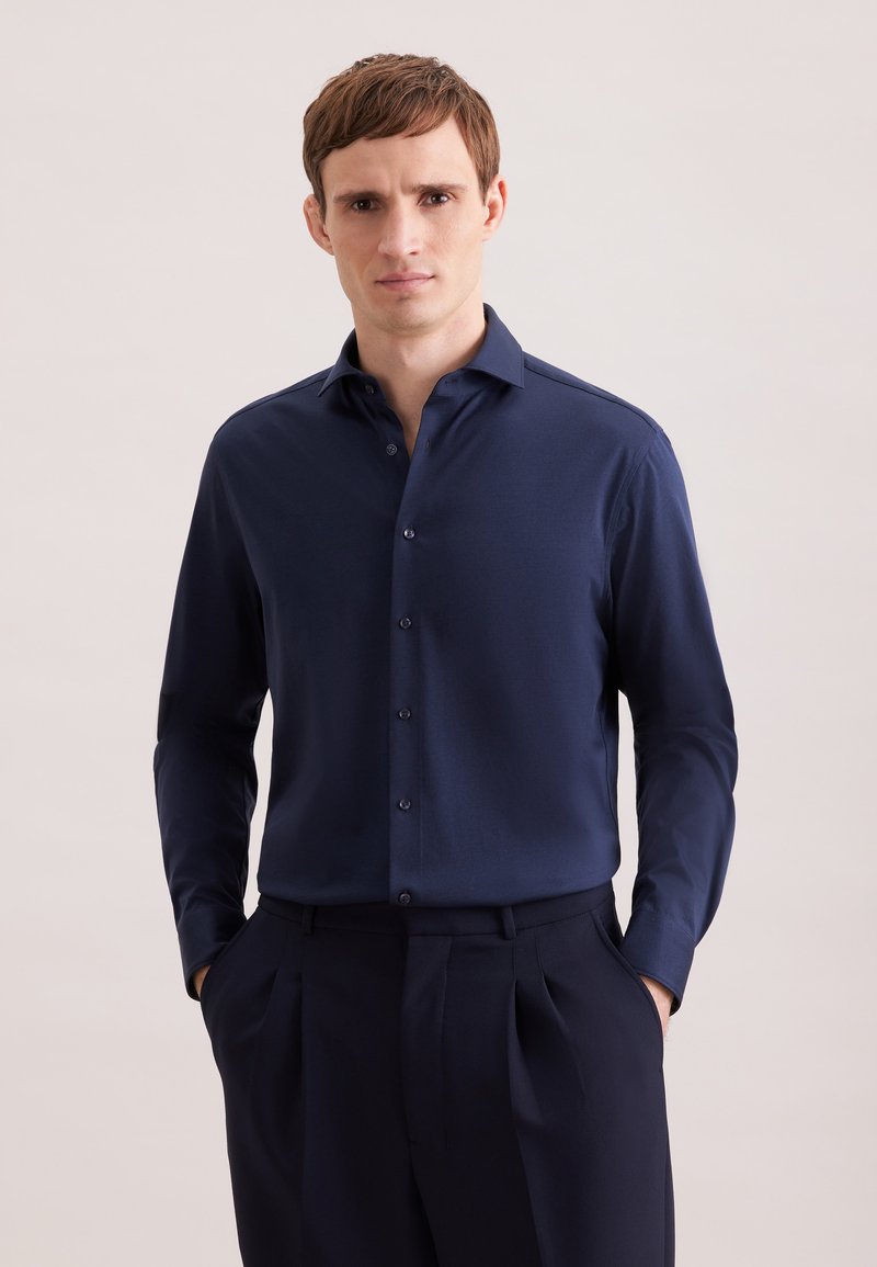 Homme portant une chemise boutonnée bleu foncé à manches longues et un pantalon sombre, debout les mains dans les poches devant un fond uni.