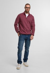 Burgundy plüss pulóver cipzáros gallérral, kék farmernadrággal és bézs sportcipőkkel párosítva. A modellen világos ing és szemüveg található.