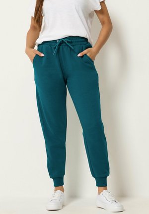 Personne portant un pantalon de jogging bleu canard avec des poignets élastiques, un t-shirt blanc, des baskets blanches, debout les mains dans les poches sur un fond uni.