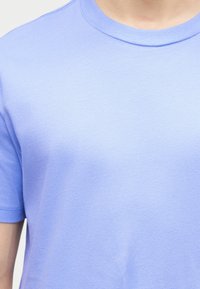 Κλειστό πλάνο ενός ατόμου που φοράει μια απλή, ανοιχτό μπλε κοντομάνικη μπλούζα με στρογγυλή λαιμόκοψη, εμφανίζοντας το άνω μέρος του στήθους και του λαιμού.