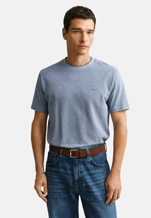 Mann trägt ein hellblaues kurzärmeliges T-Shirt, blaue Jeans mit braunem Gürtel und ein silbernes Armband am linken Handgelenk.