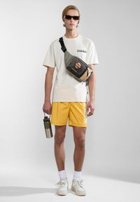 Groene en zwarte schoudertas met mesh-pocket, gele shorts met witte letters, wit T-shirt, zonnebril en een zilveren waterfles.