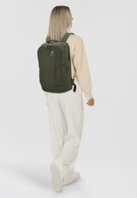 Olivgrüner Rucksack mit rechteckiger Form, ausgestattet mit einer vorderen Reißverschlusstasche, verstellbaren gepolsterten Trägern und einem Logo auf der Oberseite.