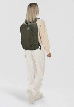 Mochila verde oliva con forma rectangular, que cuenta con un bolsillo con cremallera en el frente, correas ajustables y acolchadas, y un logo en la parte superior.