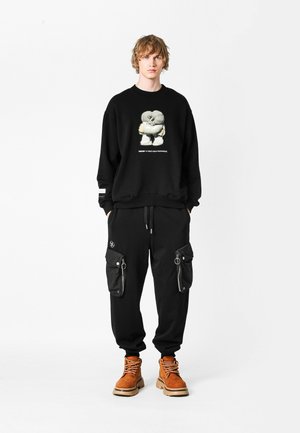 Sudadera negra de gran tamaño con diseño gráfico, combinada con pantalones cargo negros con bolsillos con cremallera y botas robustas marrones.