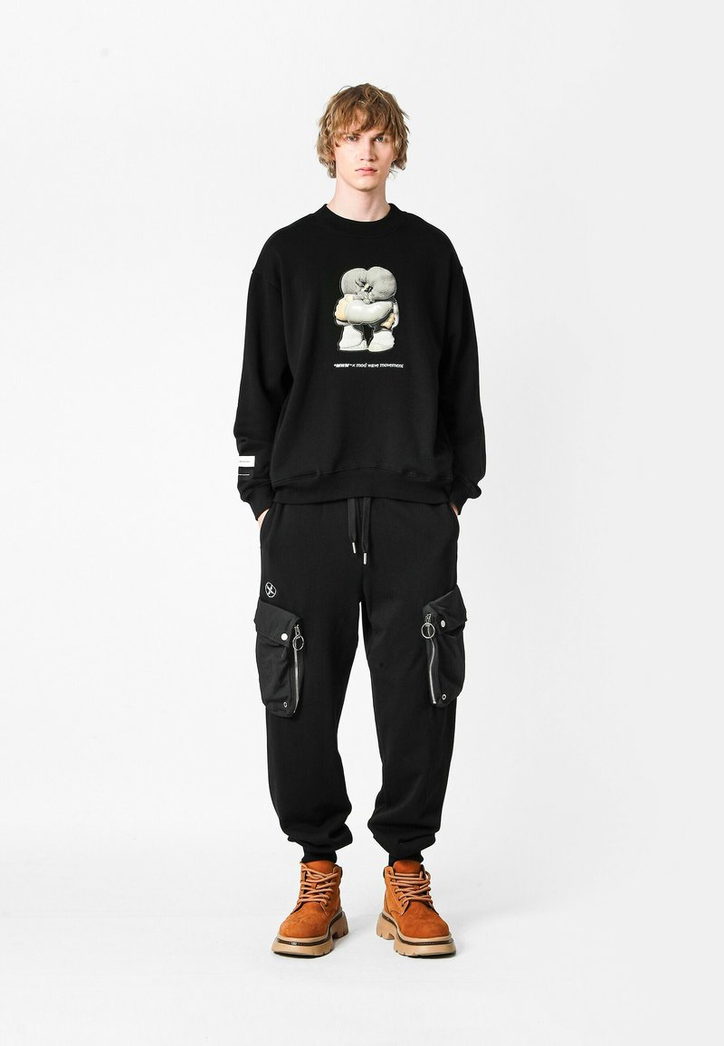 Sudadera negra de gran tamaño con diseño gráfico, combinada con pantalones cargo negros con bolsillos con cremallera y botas robustas marrones.