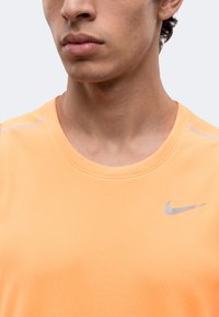 Jeune homme portant un t-shirt de sport Nike orange clair avec un logo Nike argenté sur la poitrine, vu des épaules vers le haut sur un fond uni.