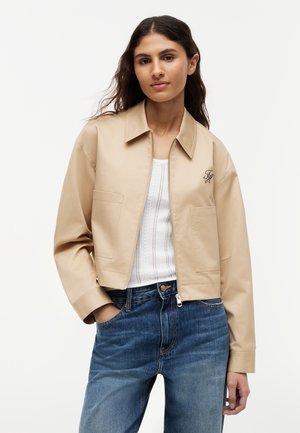 Mujer joven con chaqueta beige con cremallera sobre una camiseta de punto blanca y vaqueros azules, de pie con una mano en el bolsillo.