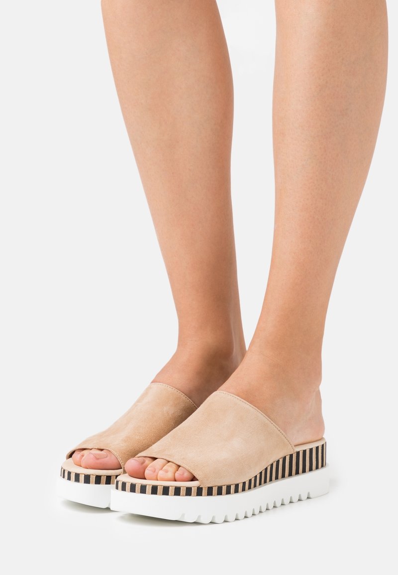 Pieds portant des sandales beige à bout ouvert, sans bride, avec une semelle intermédiaire rayée et une épaisse semelle blanche cannelée, posés sur un fond clair uni.
