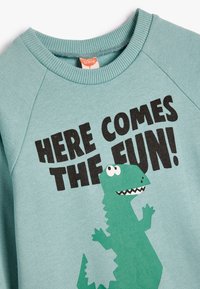 Sudadera azul claro con un gráfico de un dinosaurio verde y detalles en blanco y negro. El texto en negro dice "¡AQUÍ VIENE LA DIVERSIÓN!"